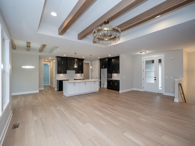 kitchen featuring a raised ceiling, a notable chandelier, an island with sink, light hardwood / wood-style floors, and decorative backsplash