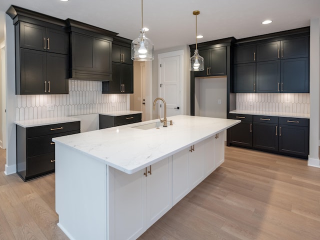 kitchen with light stone countertops, an island with sink, decorative light fixtures, decorative backsplash, and light wood-type flooring