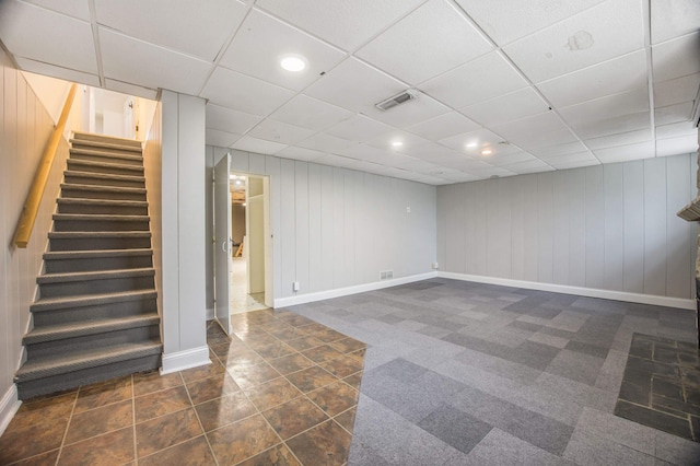 basement featuring a paneled ceiling, wooden walls, and dark colored carpet