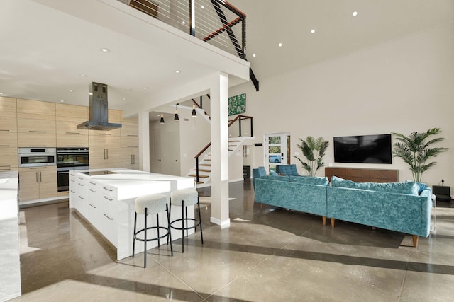 kitchen featuring light brown cabinets, wall chimney exhaust hood, a high ceiling, a kitchen island with sink, and black electric stovetop