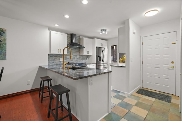 kitchen with kitchen peninsula, a kitchen breakfast bar, wall chimney exhaust hood, stainless steel fridge with ice dispenser, and white cabinetry