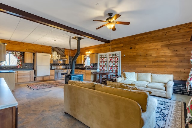 living room with ceiling fan with notable chandelier, wooden walls, sink, beam ceiling, and a wood stove