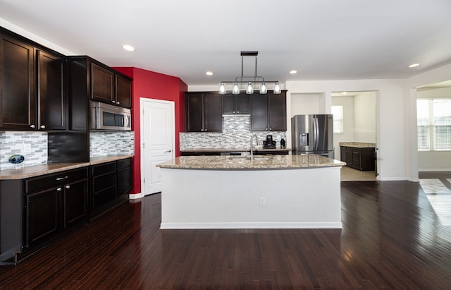 kitchen with dark wood-type flooring, an island with sink, decorative light fixtures, decorative backsplash, and appliances with stainless steel finishes