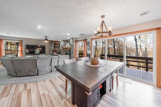 dining room with a stone fireplace, ceiling fan with notable chandelier, light hardwood / wood-style floors, and a textured ceiling