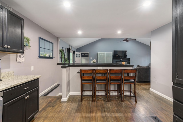 kitchen with ceiling fan, dark wood-type flooring, light stone counters, vaulted ceiling, and a breakfast bar