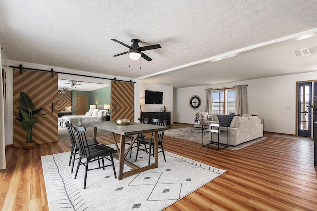 dining space featuring a textured ceiling, a barn door, wood finished floors, visible vents, and a ceiling fan