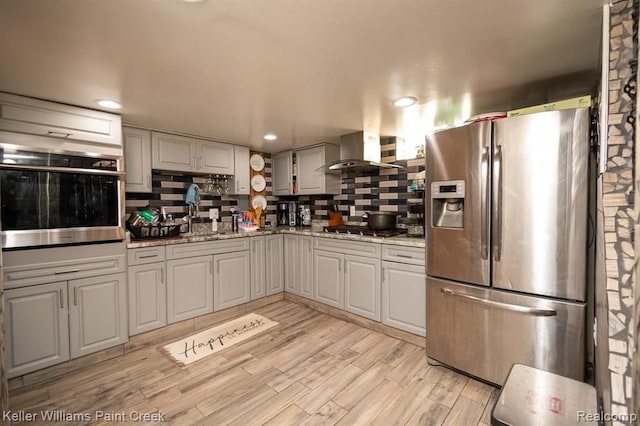 kitchen featuring backsplash, wall chimney range hood, light hardwood / wood-style flooring, light stone countertops, and stainless steel appliances