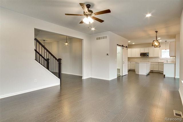 unfurnished living room with a barn door, ceiling fan, and dark hardwood / wood-style flooring