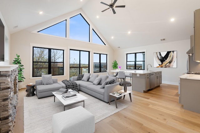 living room featuring ceiling fan, sink, a stone fireplace, high vaulted ceiling, and light wood-type flooring