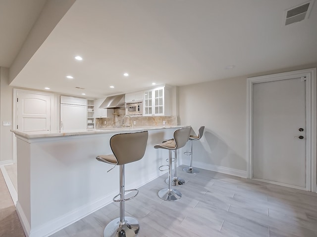 kitchen with decorative backsplash, kitchen peninsula, wall chimney exhaust hood, built in appliances, and white cabinets