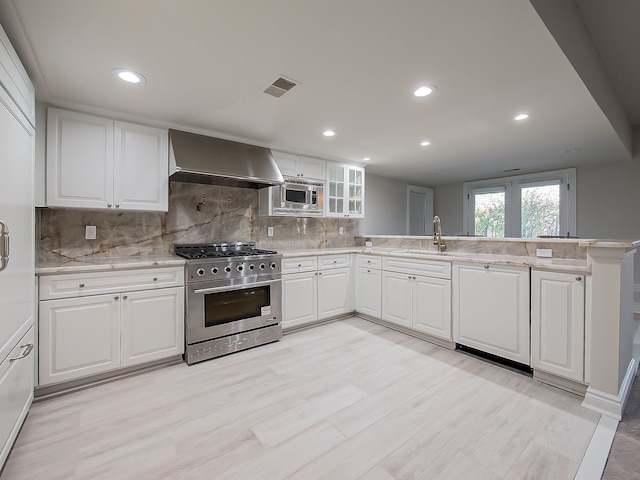 kitchen with kitchen peninsula, stainless steel appliances, sink, wall chimney range hood, and white cabinets