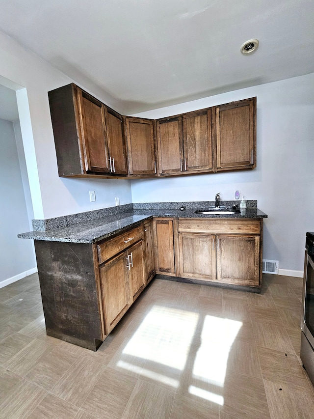 kitchen with kitchen peninsula, dark brown cabinetry, dark stone countertops, and sink