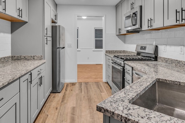 kitchen with gray cabinetry, light hardwood / wood-style flooring, decorative backsplash, light stone countertops, and appliances with stainless steel finishes