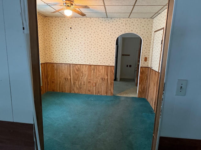 spare room with dark colored carpet, a drop ceiling, wooden walls, and ceiling fan