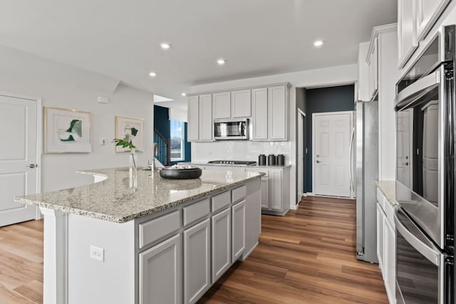 kitchen featuring dark wood-style floors, stainless steel appliances, an island with sink, and light stone countertops