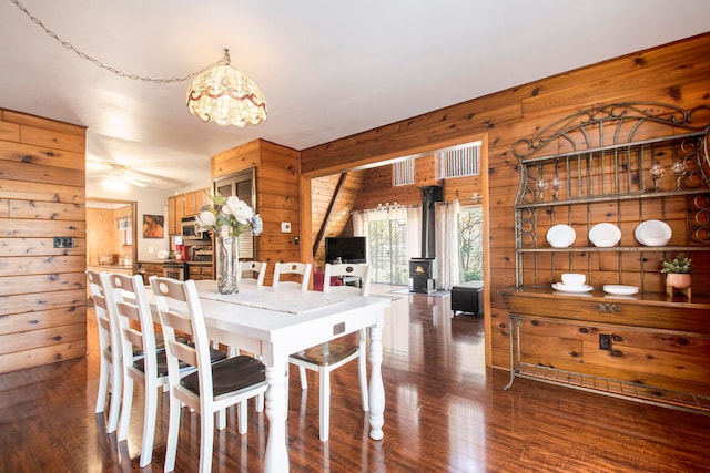 dining area with dark hardwood / wood-style floors, a wood stove, ceiling fan, and wood walls