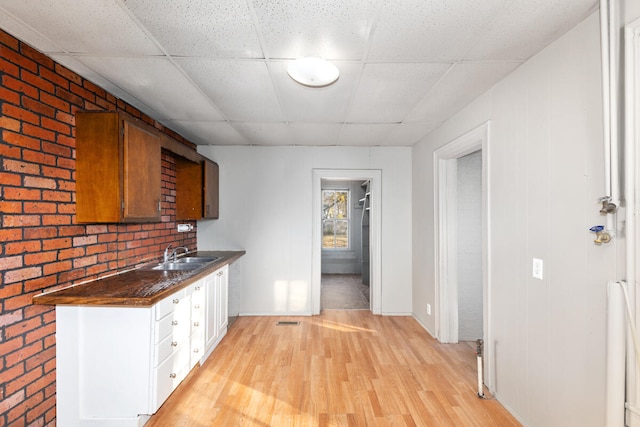 kitchen with a drop ceiling, sink, light hardwood / wood-style floors, white cabinetry, and brick wall