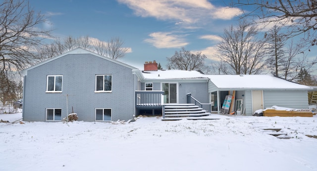 snow covered house with a deck, brick siding, and a chimney