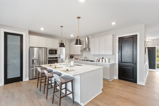 kitchen with white cabinetry, hanging light fixtures, stainless steel appliances, wall chimney range hood, and a kitchen island with sink