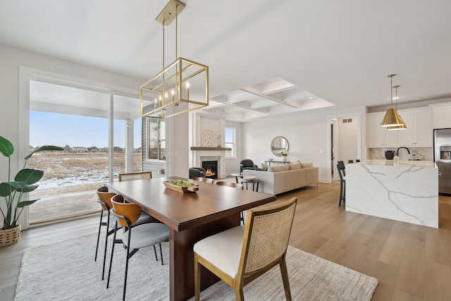 dining area featuring a warm lit fireplace, baseboards, coffered ceiling, light wood-type flooring, and beam ceiling