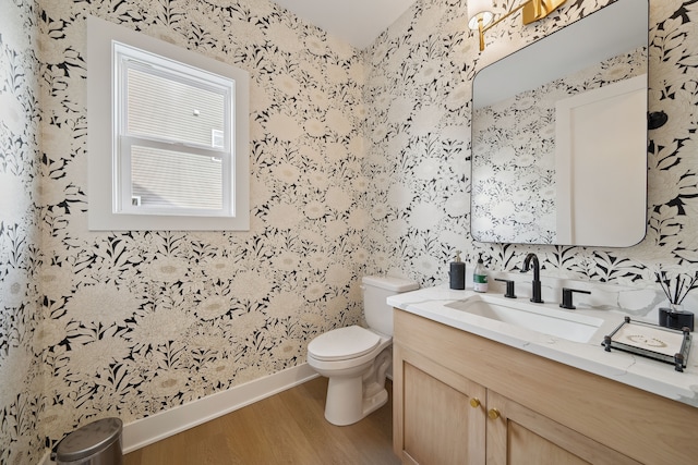 half bathroom featuring wallpapered walls, baseboards, toilet, wood finished floors, and vanity