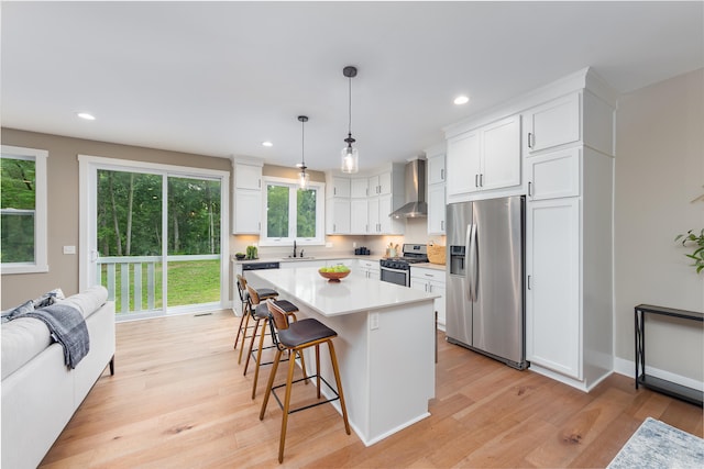 kitchen featuring appliances with stainless steel finishes, wall chimney range hood, light hardwood / wood-style flooring, white cabinets, and a kitchen island