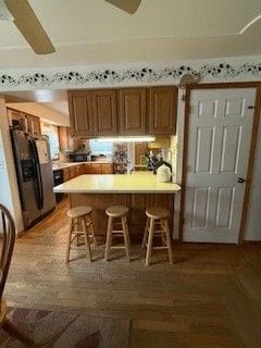 kitchen with kitchen peninsula, ceiling fan, wood-type flooring, stainless steel fridge with ice dispenser, and a breakfast bar area