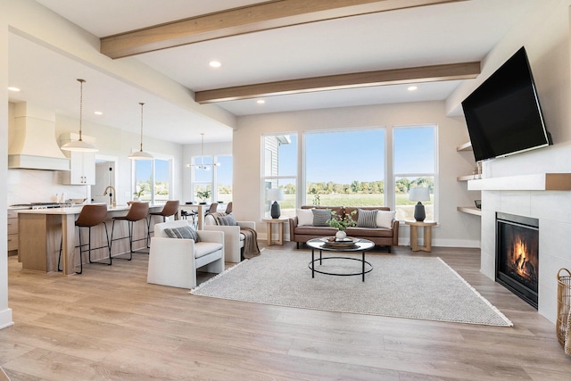 living room featuring a tiled fireplace, sink, beamed ceiling, light hardwood / wood-style floors, and a chandelier