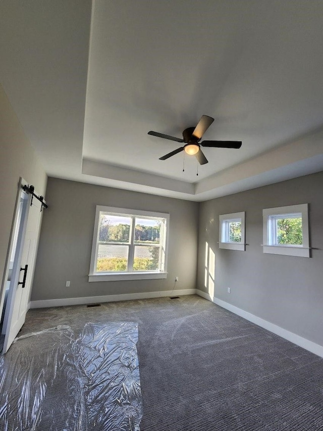 empty room featuring dark colored carpet, ceiling fan, a barn door, and a healthy amount of sunlight