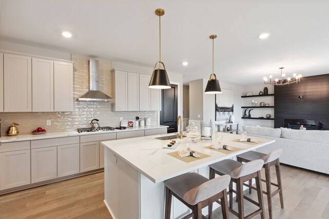 kitchen featuring an island with sink, decorative light fixtures, light hardwood / wood-style flooring, and wall chimney range hood