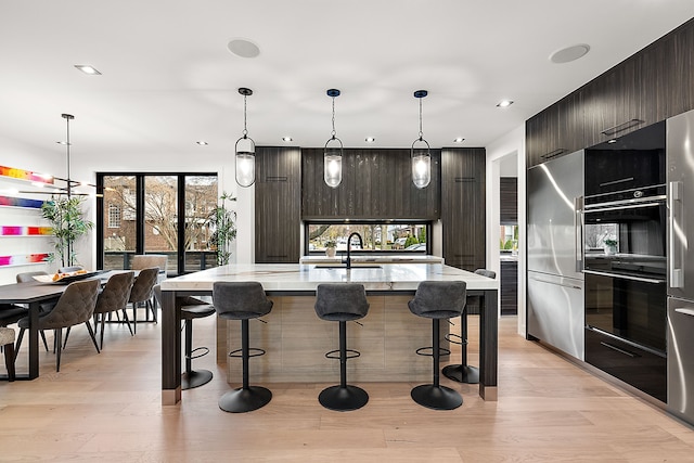 kitchen with high end fridge, light hardwood / wood-style flooring, decorative light fixtures, dark brown cabinets, and a center island with sink