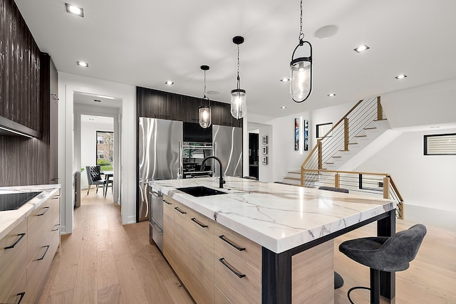 kitchen featuring light stone countertops, light wood-type flooring, an island with sink, decorative light fixtures, and stainless steel refrigerator