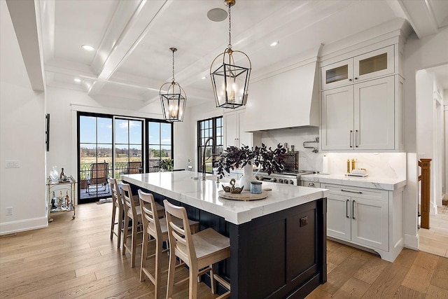 kitchen with beam ceiling, decorative light fixtures, white cabinets, light hardwood / wood-style floors, and an island with sink