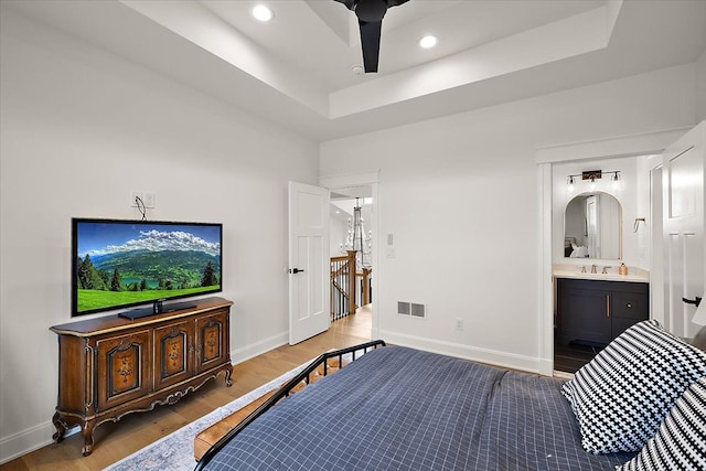 bedroom with sink, ensuite bath, ceiling fan, light wood-type flooring, and a tray ceiling