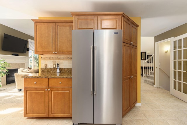 kitchen with high end refrigerator, decorative backsplash, dark stone countertops, a fireplace, and light tile patterned flooring