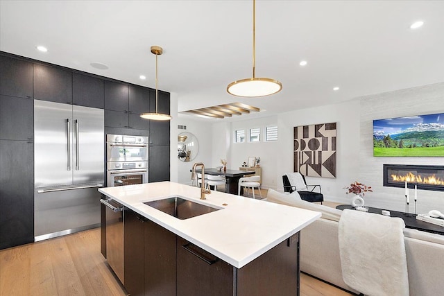 kitchen with appliances with stainless steel finishes, a kitchen island with sink, sink, decorative light fixtures, and light hardwood / wood-style floors