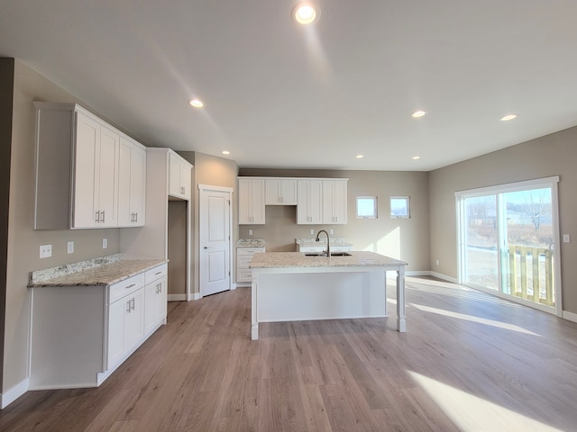 kitchen with a center island with sink, white cabinets, sink, light hardwood / wood-style flooring, and light stone countertops