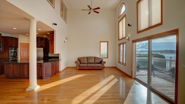 living room featuring a towering ceiling, decorative columns, ceiling fan, a water view, and light hardwood / wood-style floors