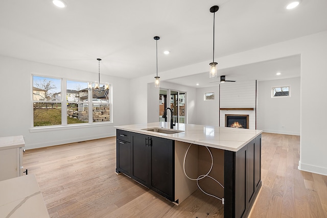 kitchen with ceiling fan with notable chandelier, sink, light wood-type flooring, an island with sink, and a large fireplace