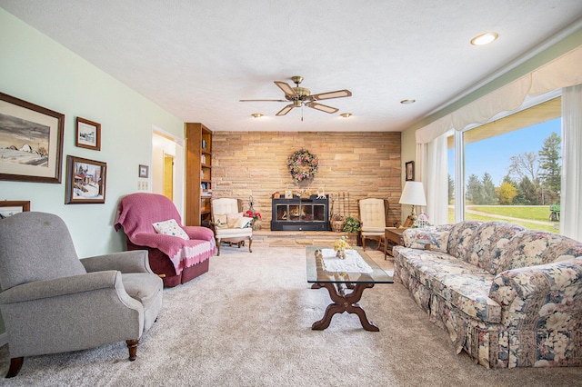 carpeted living room featuring ceiling fan, wood walls, a stone fireplace, and a textured ceiling