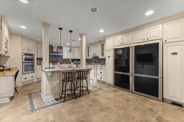 kitchen featuring pendant lighting, a kitchen island with sink, wall chimney exhaust hood, double oven, and light stone counters