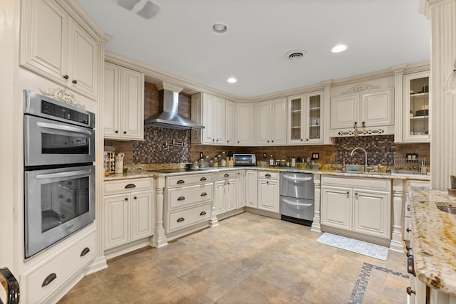 kitchen featuring light stone countertops, tasteful backsplash, wall chimney exhaust hood, double oven, and cream cabinetry