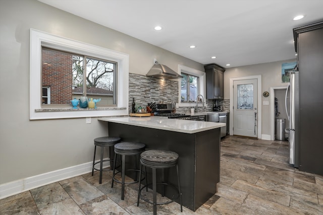 kitchen featuring a kitchen bar, appliances with stainless steel finishes, kitchen peninsula, tasteful backsplash, and wall chimney exhaust hood