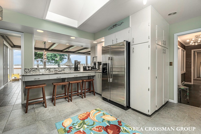 kitchen featuring stainless steel refrigerator with ice dispenser, backsplash, a breakfast bar, beam ceiling, and white cabinetry