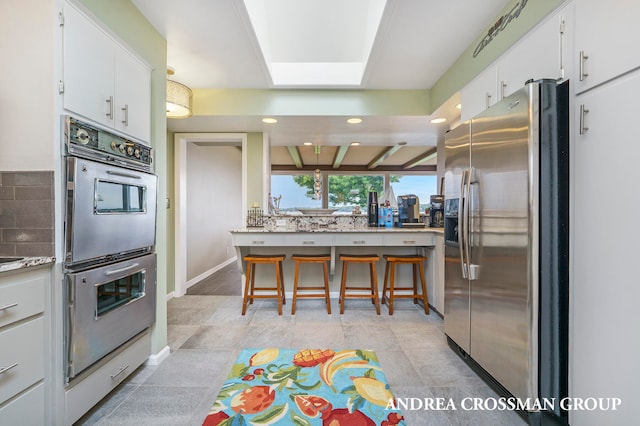 kitchen featuring decorative backsplash, light tile patterned floors, beam ceiling, white cabinetry, and stainless steel appliances