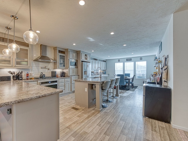 kitchen featuring appliances with stainless steel finishes, wall chimney exhaust hood, decorative light fixtures, light hardwood / wood-style floors, and a breakfast bar area