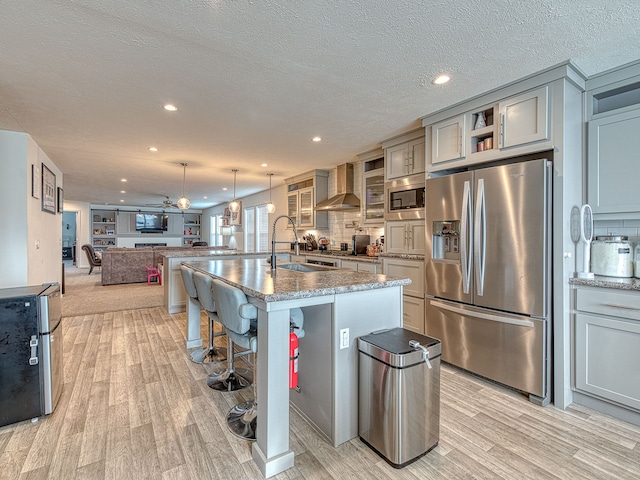 kitchen featuring sink, wall chimney exhaust hood, stainless steel appliances, light stone counters, and a kitchen island with sink