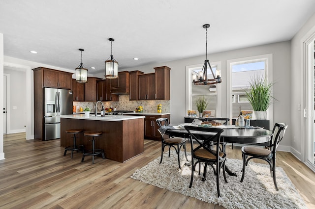 kitchen with decorative light fixtures, stainless steel refrigerator with ice dispenser, light countertops, a kitchen island with sink, and dark brown cabinetry