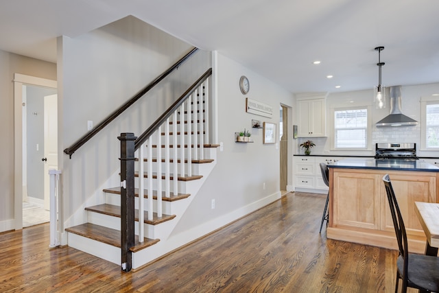 kitchen featuring dark wood-type flooring, wall chimney range hood, light brown cabinets, decorative light fixtures, and stainless steel range oven