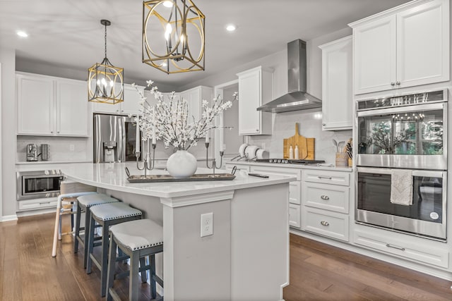 kitchen featuring pendant lighting, a center island with sink, wall chimney exhaust hood, white cabinetry, and stainless steel appliances